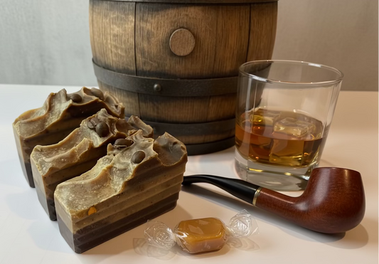 Three pieces of brown soap, a wooden barrel, a glass with amber liquid, and a wooden pipe on a white surface.
