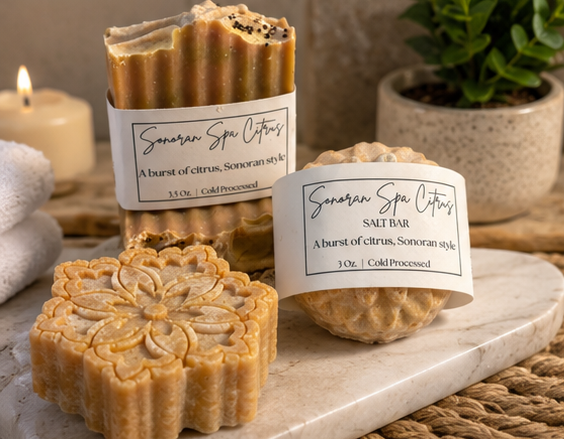 Three soap bars with labels on a marble surface, with a candle and plant in the background.
